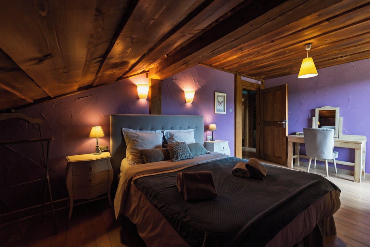 Ein Schlafzimmer mit Holzbalkendecke, einem Bett mit vielen Kissen, Nachttischlampen und einem Schreibtisch mit Stuhl.