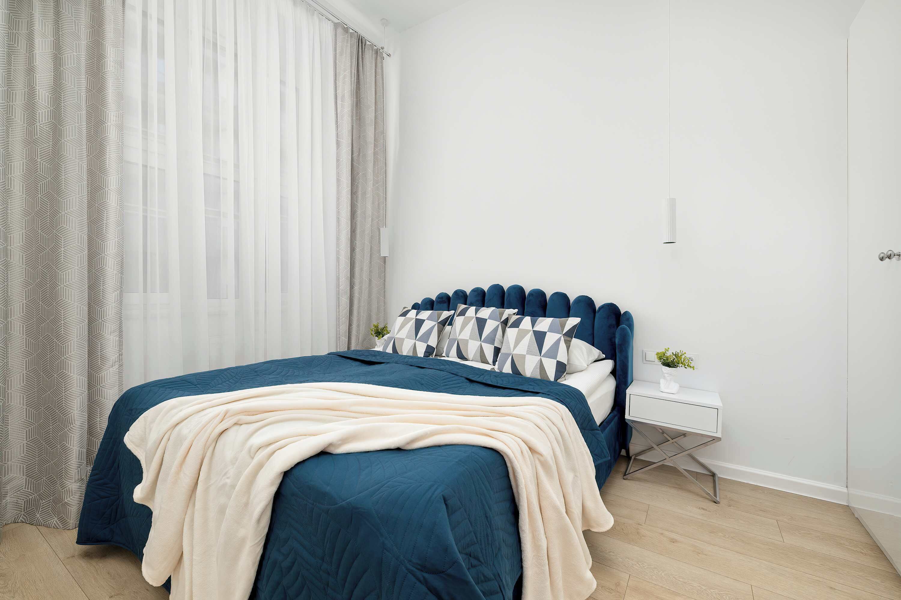 An elegant bedroom with a large double bed, dressed in a stylish navy blue bedspread and a thick, cream-colored blanket. Light-colored walls and delicate curtains create a peaceful atmosphere conducive to relaxation.