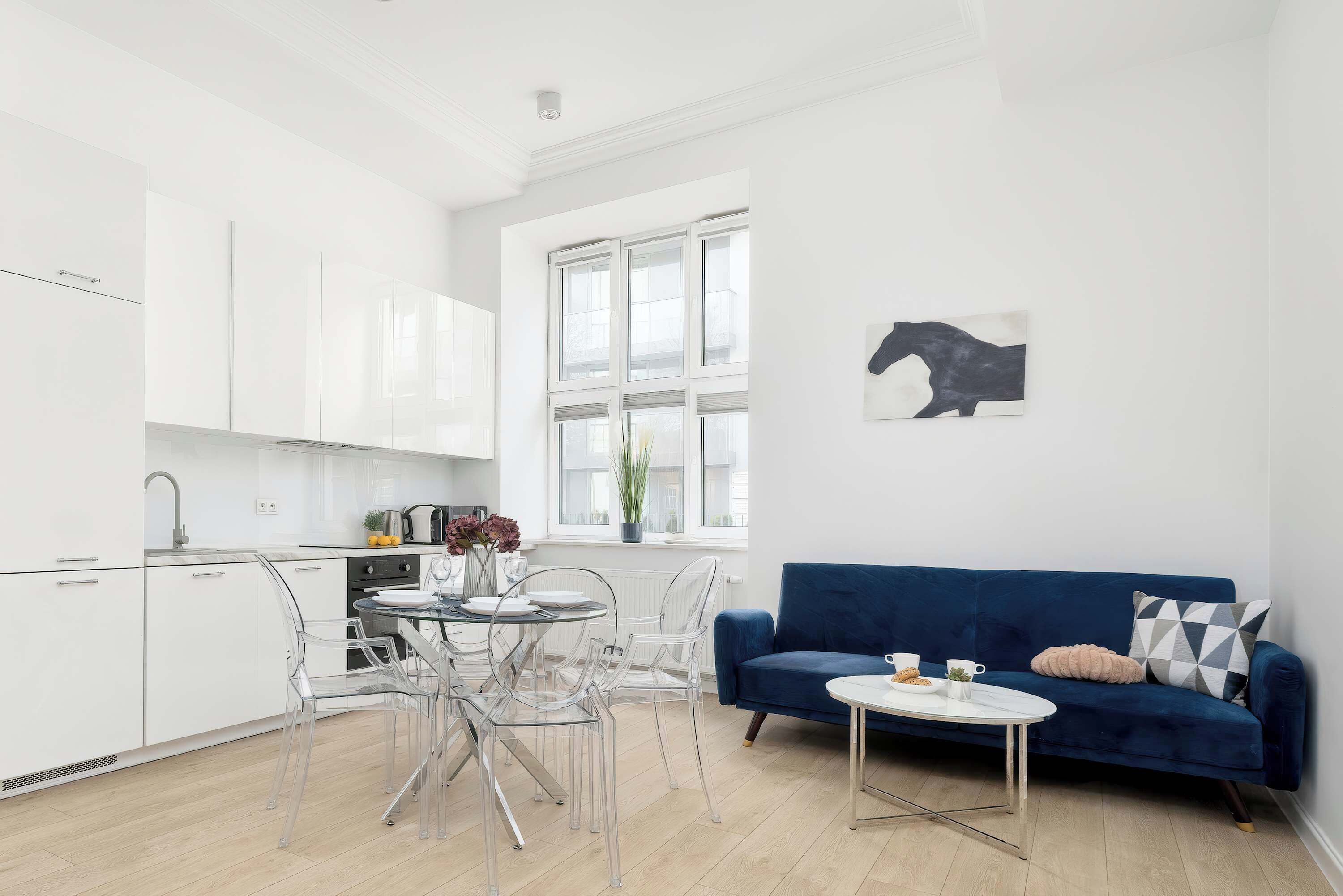 A modern kitchen with white cabinets, a dining area with clear chairs, and a blue sofa with a small table.