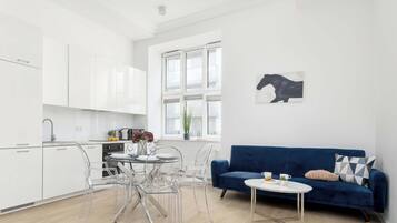A modern kitchen with white cabinets, a dining area with clear chairs, and a blue sofa with a small table.