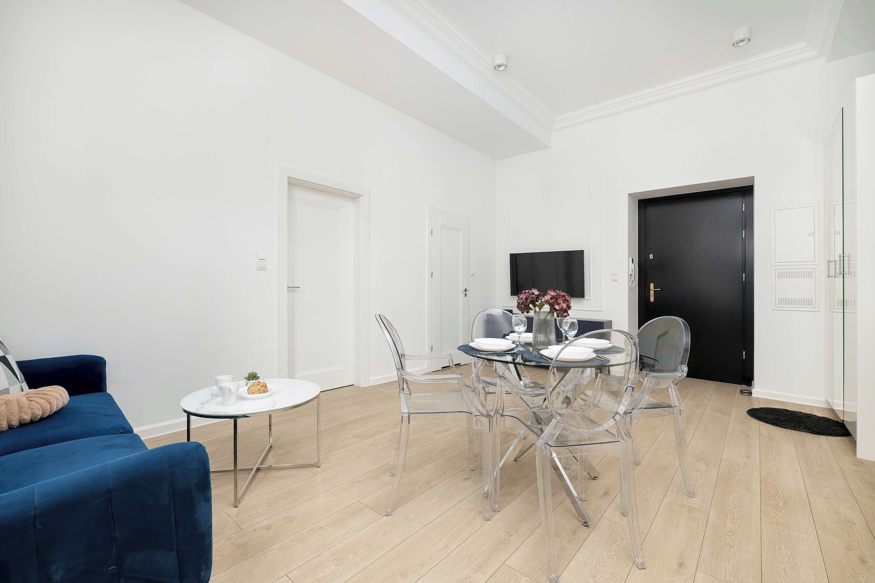 A modern living room with a blue sofa, a round dining table with clear chairs, and a black door in the background.