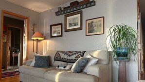 Een woonkamer met een beige bank, decoratieve kussens, een lamp, ingelijste foto's aan de muur en een potplant.