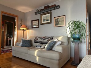 A living room with a beige sofa, patterned cushions, a lamp, framed pictures on the wall, and a potted plant. - Sunset Paradise! Pet-Friendly Soundfront Home on Kitty Hawk Bay! (Kill Devil Hills)