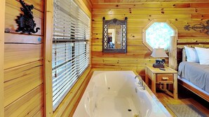 A wooden cabin interior with a large bathtub, a bed, a mirror, and a window with blinds. - Smoky Mountain View (Sevierville)