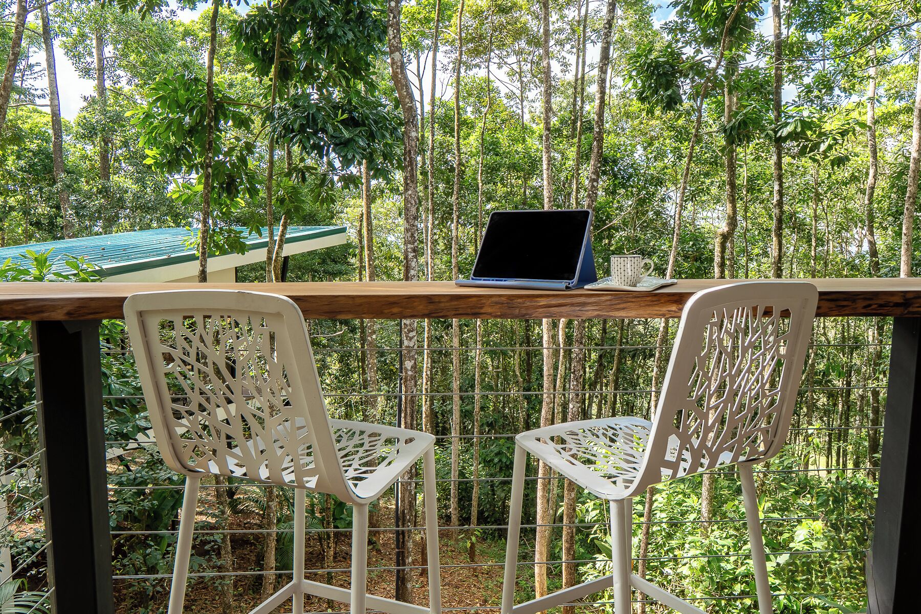 A wooden deck with a laptop and a cup on a table, surrounded by white chairs, set against a forest backdrop.