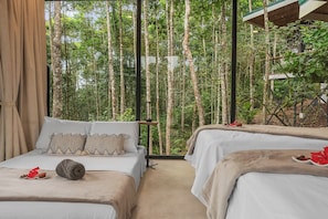 A room with two beds, a window with a view of tall trees, and a small table with a lamp.