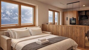 A modern bedroom with a large bed, wooden wardrobe, and a kitchen area visible through a glass partition.