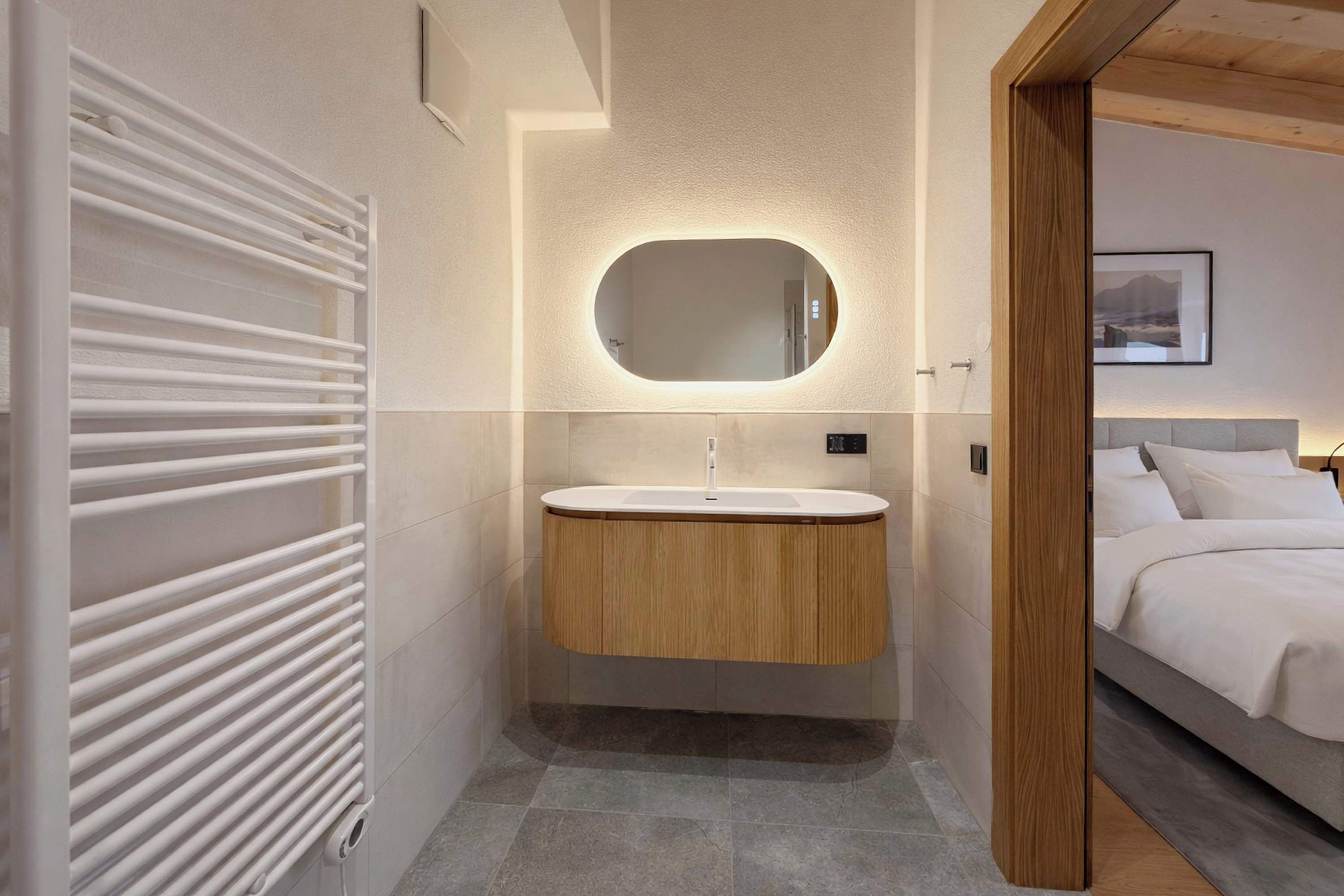 Ein modernes Badezimmer mit einem weißen Heizkörper, einem hölzernen Waschtisch, einem runden Spiegel und einem Bett mit weißer Bettwäsche.