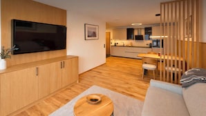 Ein modernes Wohnzimmer mit Fernseher, Holzschränken, einer Couch, einer Essecke mit Tisch und Stühlen sowie einer Küche im Hintergrund.