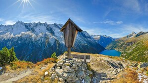 A wooden mountain hut with a stone base, surrounded by rocky terrain and a lake, with snow-capped mountains in the background.