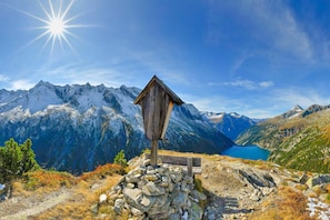 Un refuge de montagne en bois, avec une base en pierre, entouré d’un paysage rocheux et d’un lac, et des montagnes enneigées en arrière-plan.