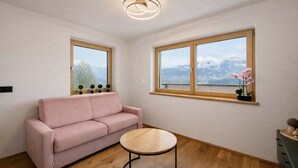 A modern living room with a pink sofa, a round wooden coffee table, and two windows with mountain views.