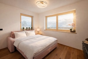 A bedroom with a bed, two windows, a bedside table, a lamp, and a view of the outdoors.