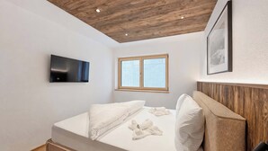 Ein modernes Schlafzimmer mit Holzboden, einem Flachbildfernseher, einem großen Fenster und einem Bett mit weißen Bettwäsche.