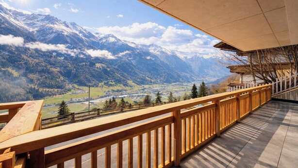 Ein Balkon mit Holzgeländer, der auf eine gebirgige Landschaft mit schneebedeckten Gipfeln und ein Tal blickt.