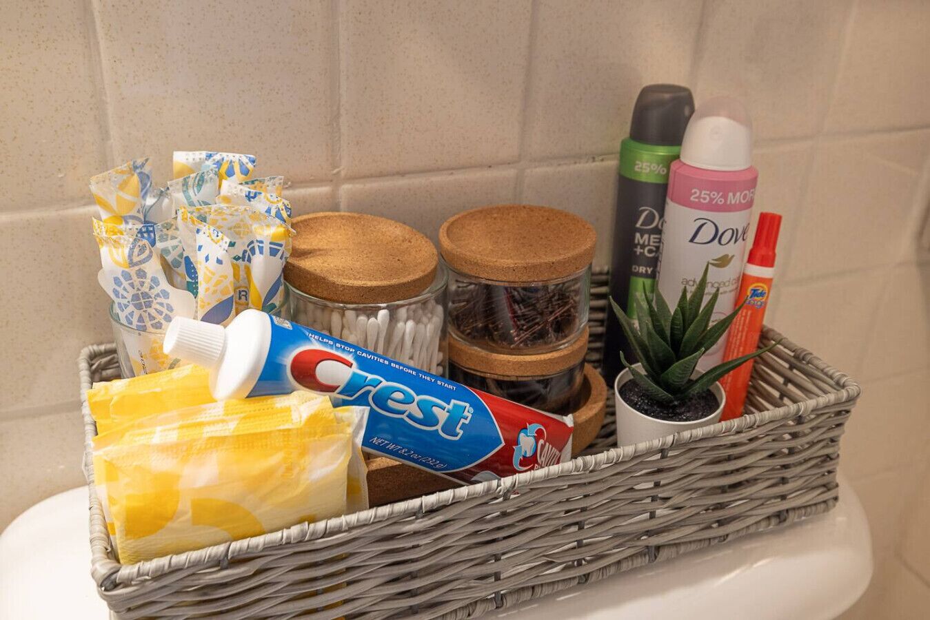 Una cesta de mimbre con artículos de higiene bucal, un tubo de pasta dental Crest, hisopos, una planta pequeña en maceta y una botella de jabón Dove.