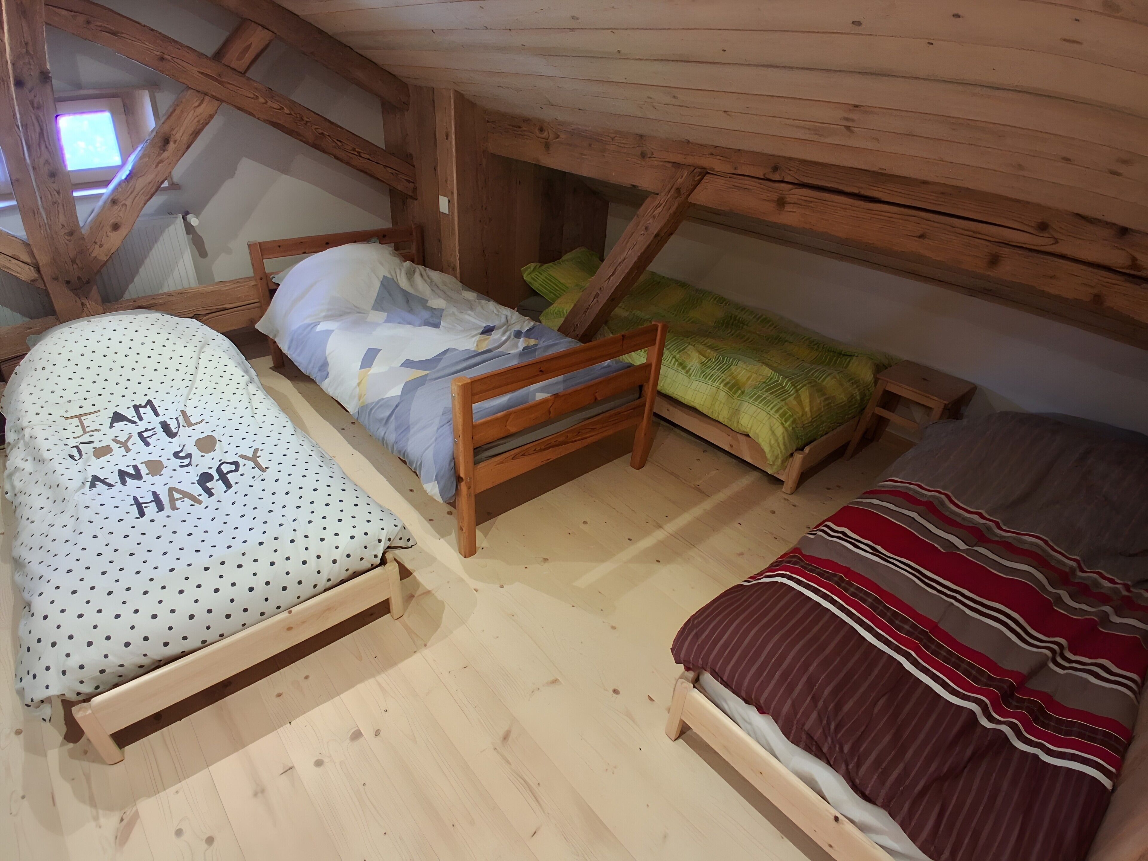 Une chambre de style « loft » avec un plafond en pente, des poutres en bois et une petite fenêtre. On y trouve trois lits avec différents types de literie, ainsi qu’une chaise en bois avec un coussin à pois.