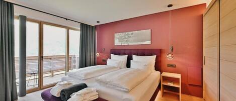 Una habitación de hotel moderna con una cama grande, un banco con toallas, una mesita de noche pequeña y un ventanal con cortinas.