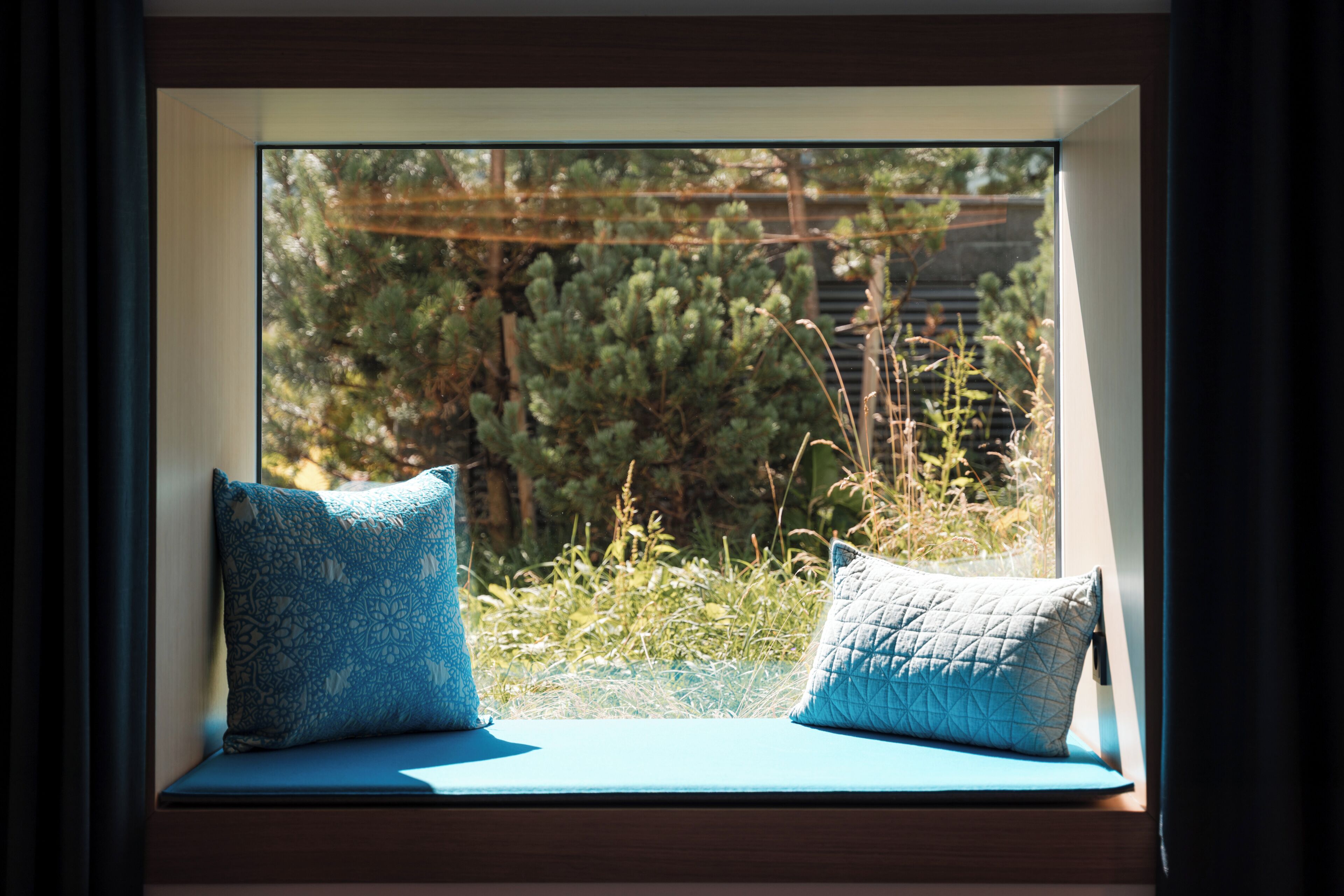 A window seat with two pillows, a view of greenery and a wooden structure outside.