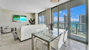 A modern living room with a glass dining table, white sofa, and a large window overlooking a cityscape and ocean. - Spacious 3-bedroom apartment Ocean View in lovely Hallandale Beach (Hallandale Beach)