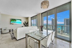 Ein modernes Wohnzimmer mit einem Glastisch, einem weißen Sofa und einem großen Fenster mit Blick auf eine Stadtlandschaft und den Ozean.