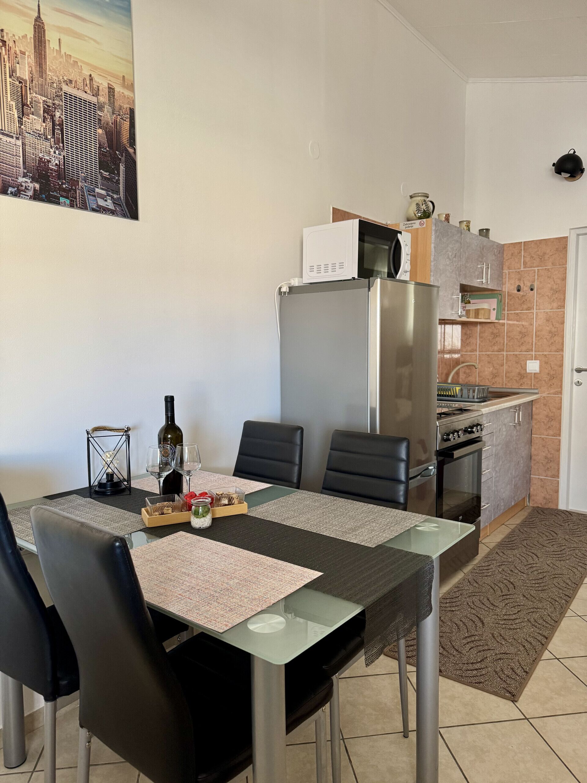 Un coin repas avec une table dressée pour quatre personnes, une cuisine équipée d’un réfrigérateur et d’un four à micro-ondes, et un tableau représentant un paysage urbain accroché au mur.