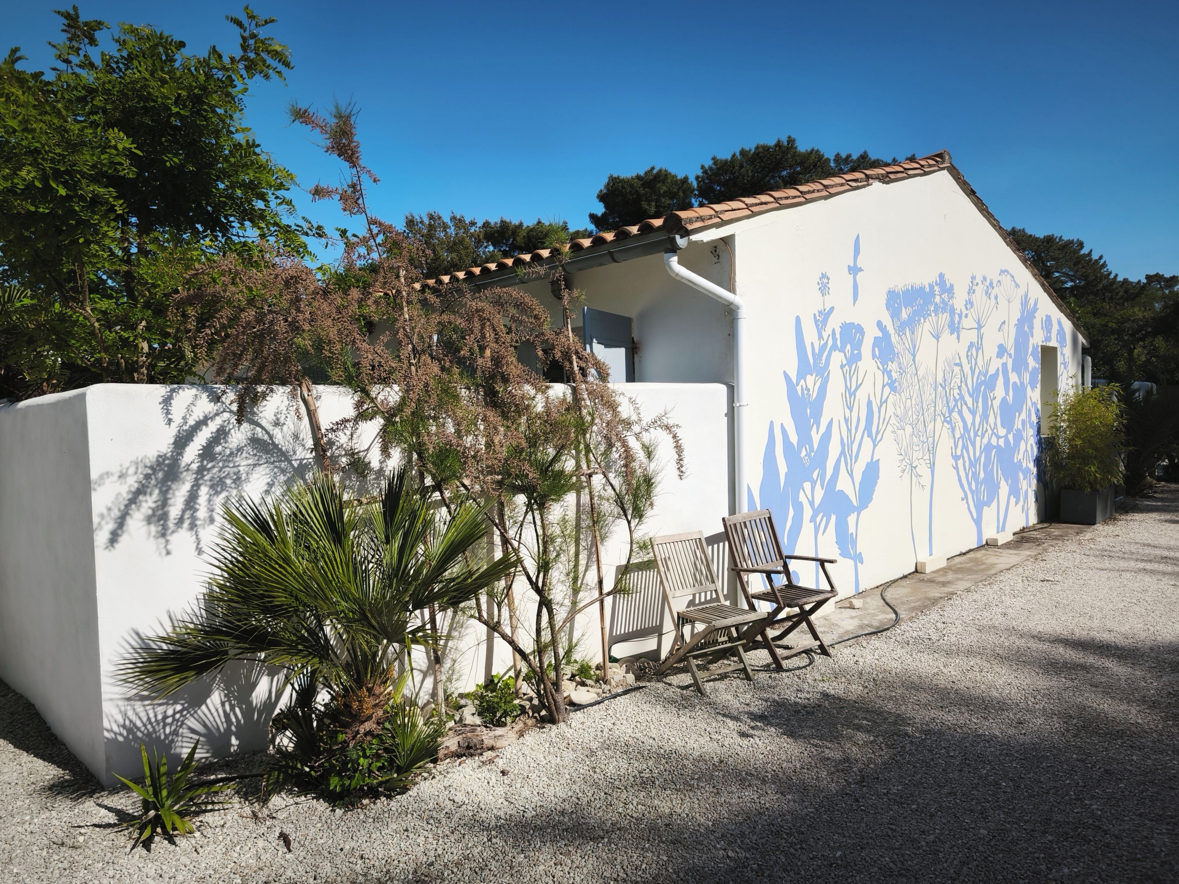 A building with a white exterior and blue floral wall art, a gravel driveway, and two wooden chairs.