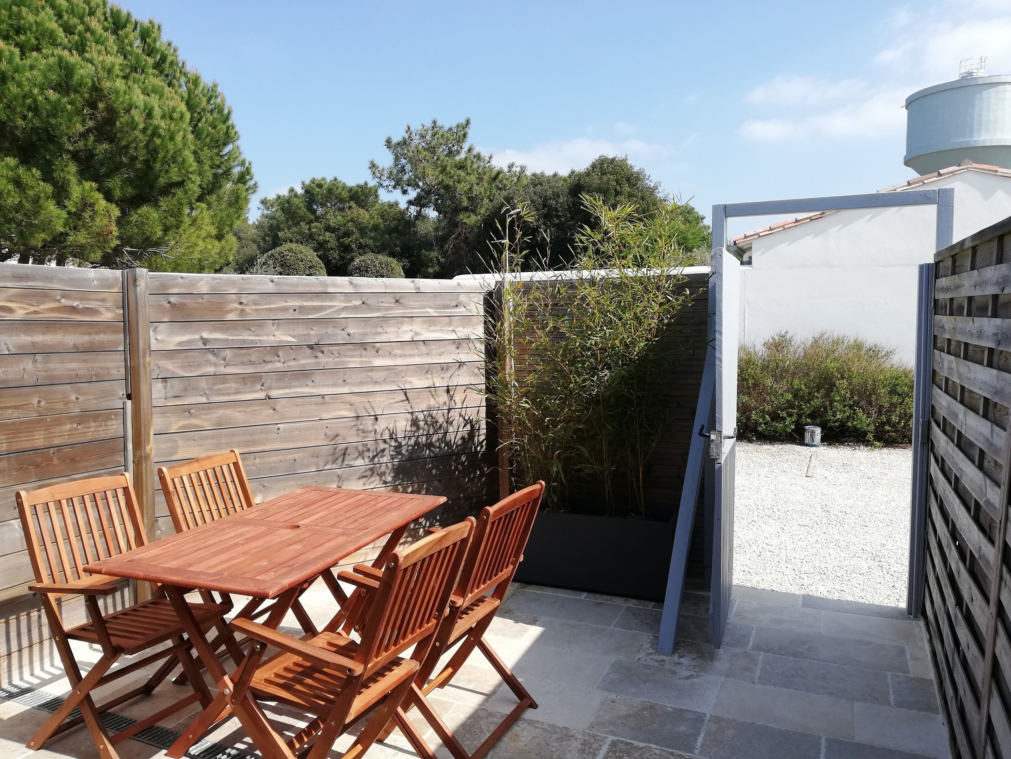 Une terrasse extérieure avec une table et des chaises en bois, une clôture en bois et un réservoir d’eau en arrière-plan.