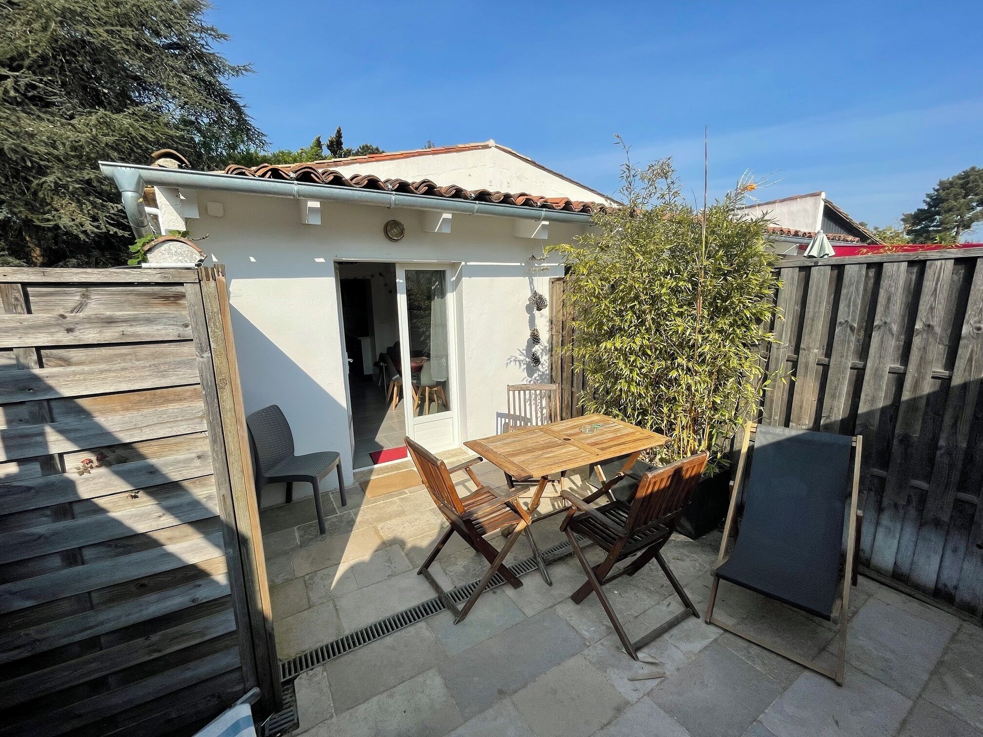 A patio with a table and chairs, a wooden fence, and a small white building with a door.