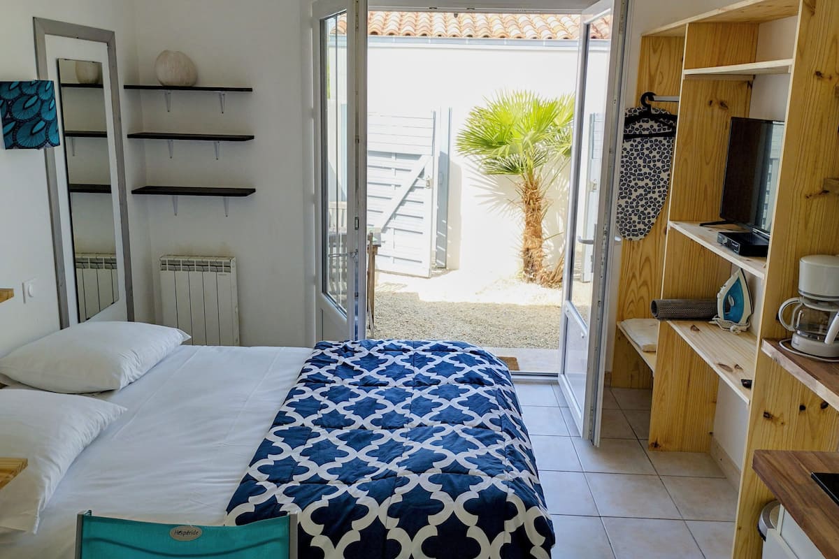 Une chambre avec un lit, une étagère en bois, un radiateur et une porte donnant sur un espace extérieur où se trouve un palmier.