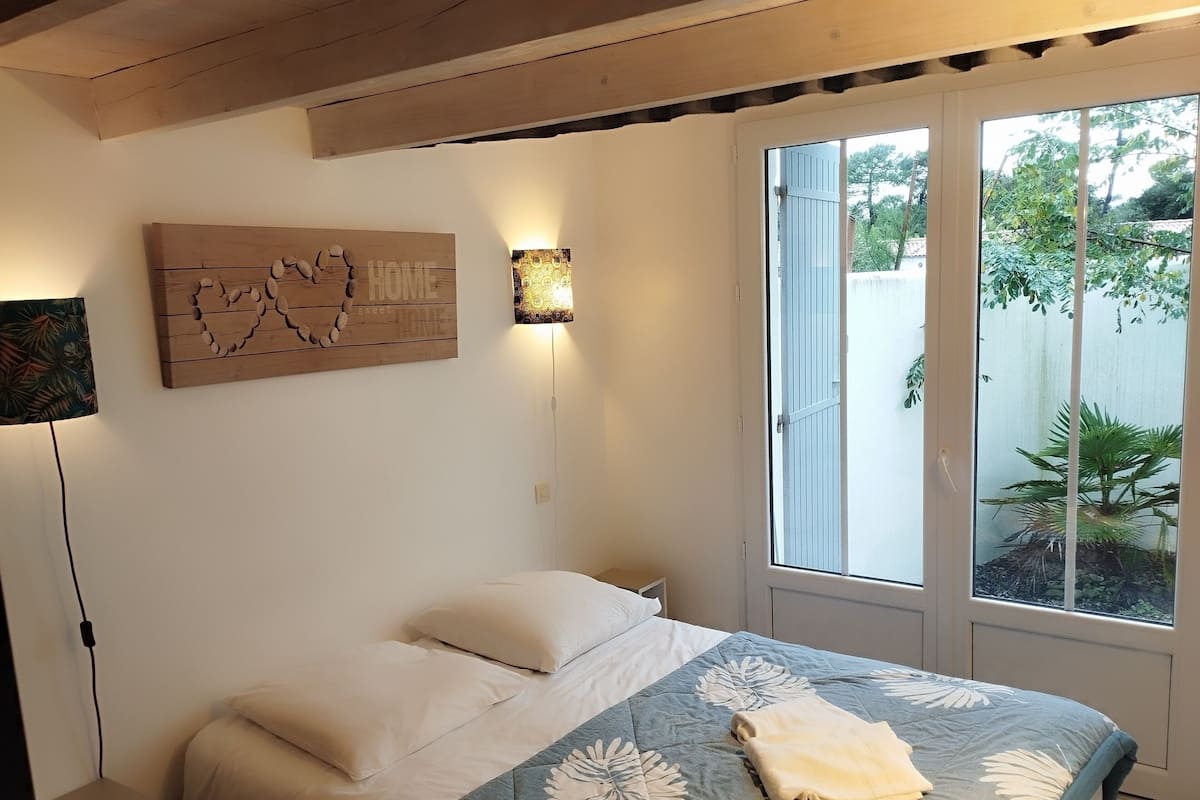 Une chambre avec un lit, une lampe fixée au mur, une fenêtre donnant sur un paysage verdoyant et une œuvre d’art en bois sur laquelle est inscrit le mot « MAISON ».