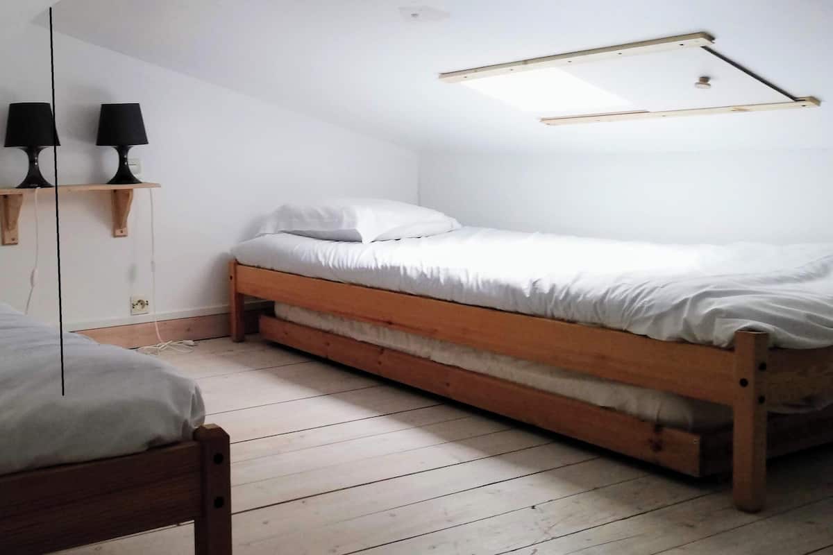 Une chambre à coucher avec un lit en bois, du linge de lit blanc, une petite étagère avec deux lampes et un plafond en pente.