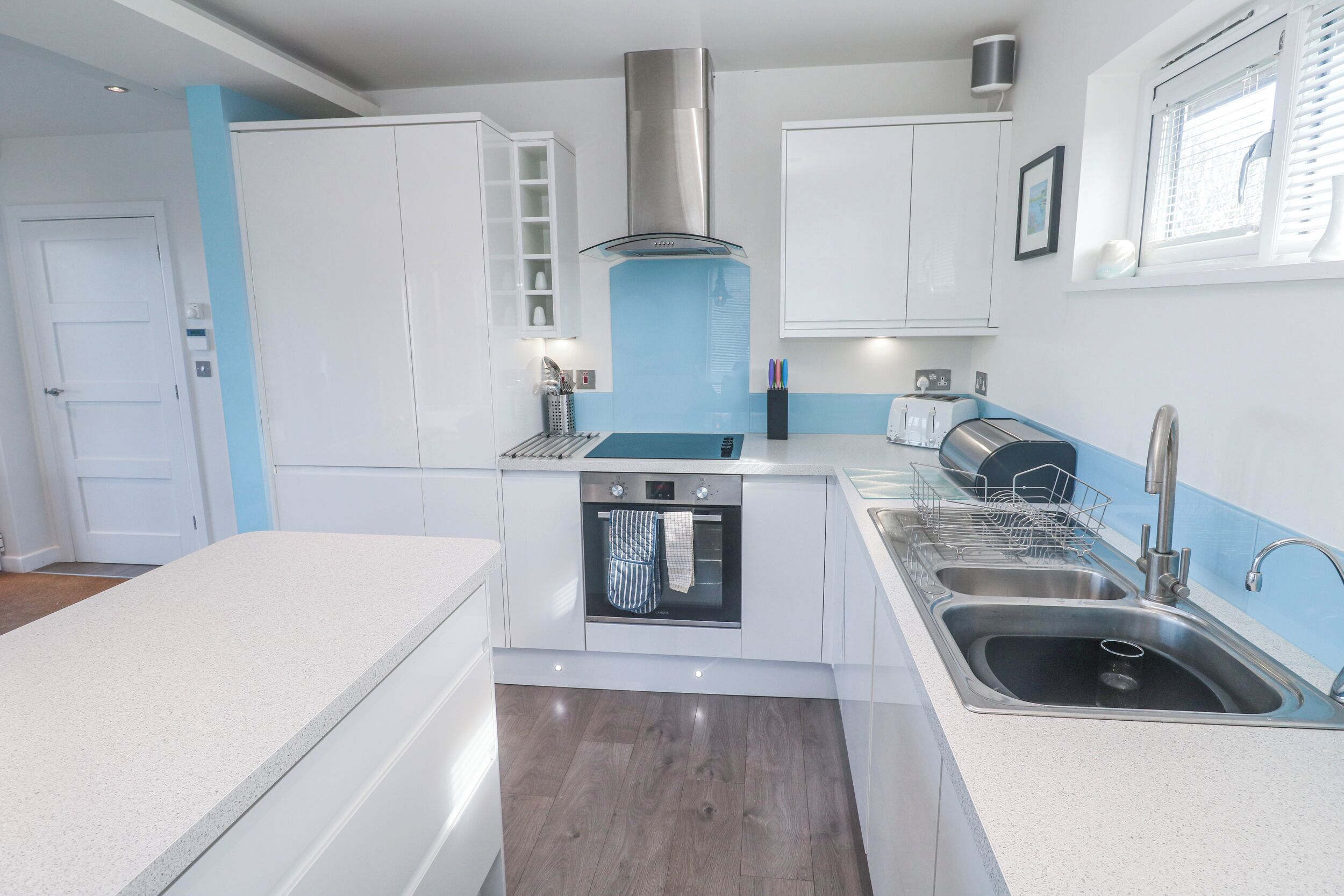 A compact kitchen with white cabinets, a stainless steel range hood, a built-in oven, a double sink, and a window with blinds.