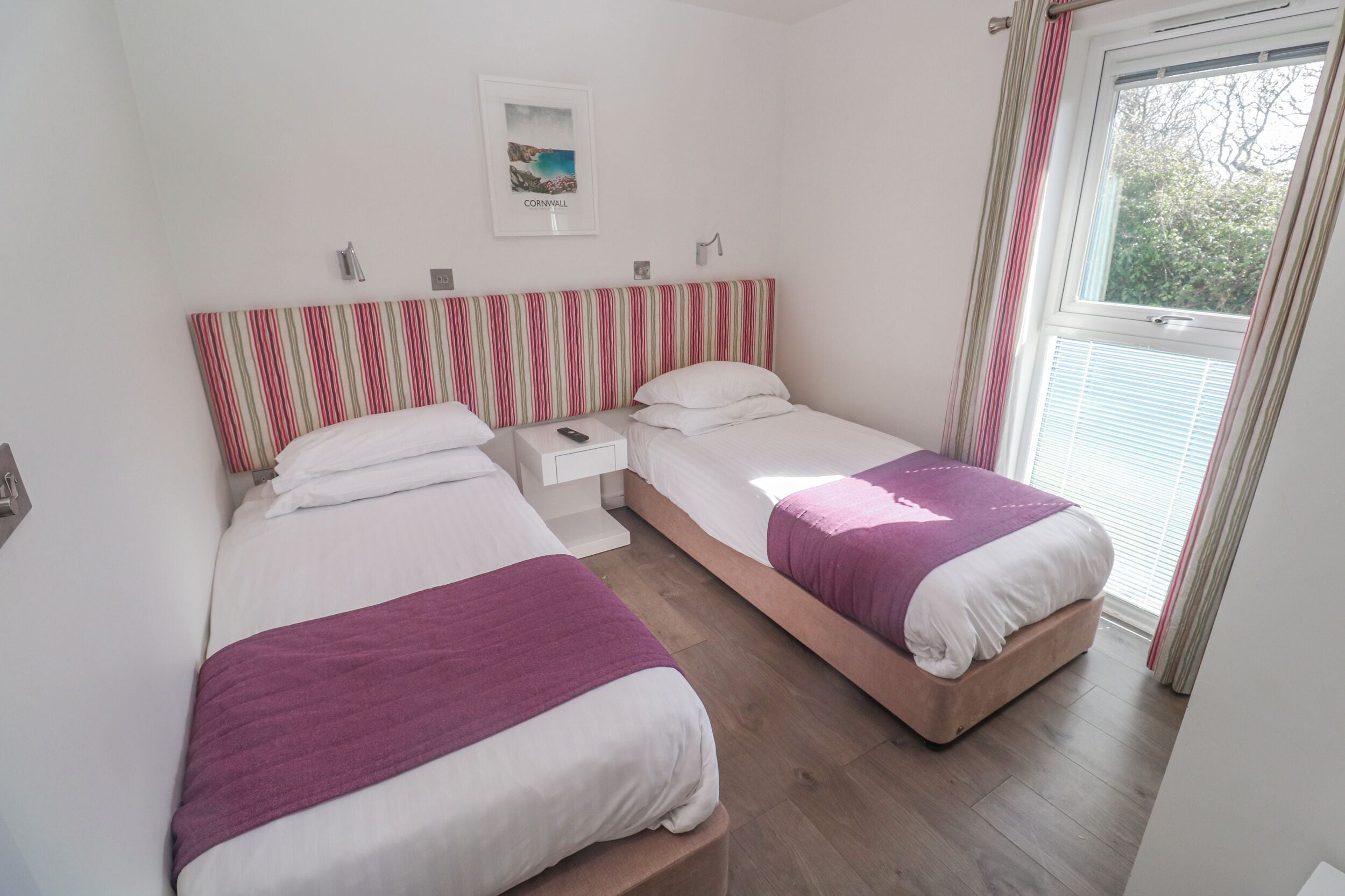 Ein Schlafzimmer mit zwei Betten, einem Nachttisch, einem gerahmten Bild an der Wand und einem Fenster mit Vorhängen.