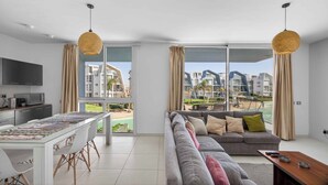 A modern living room with a sofa, dining table, and kitchen area, featuring large windows and a view of buildings. - BirdNest At Fouka Bay (El Dabaa)