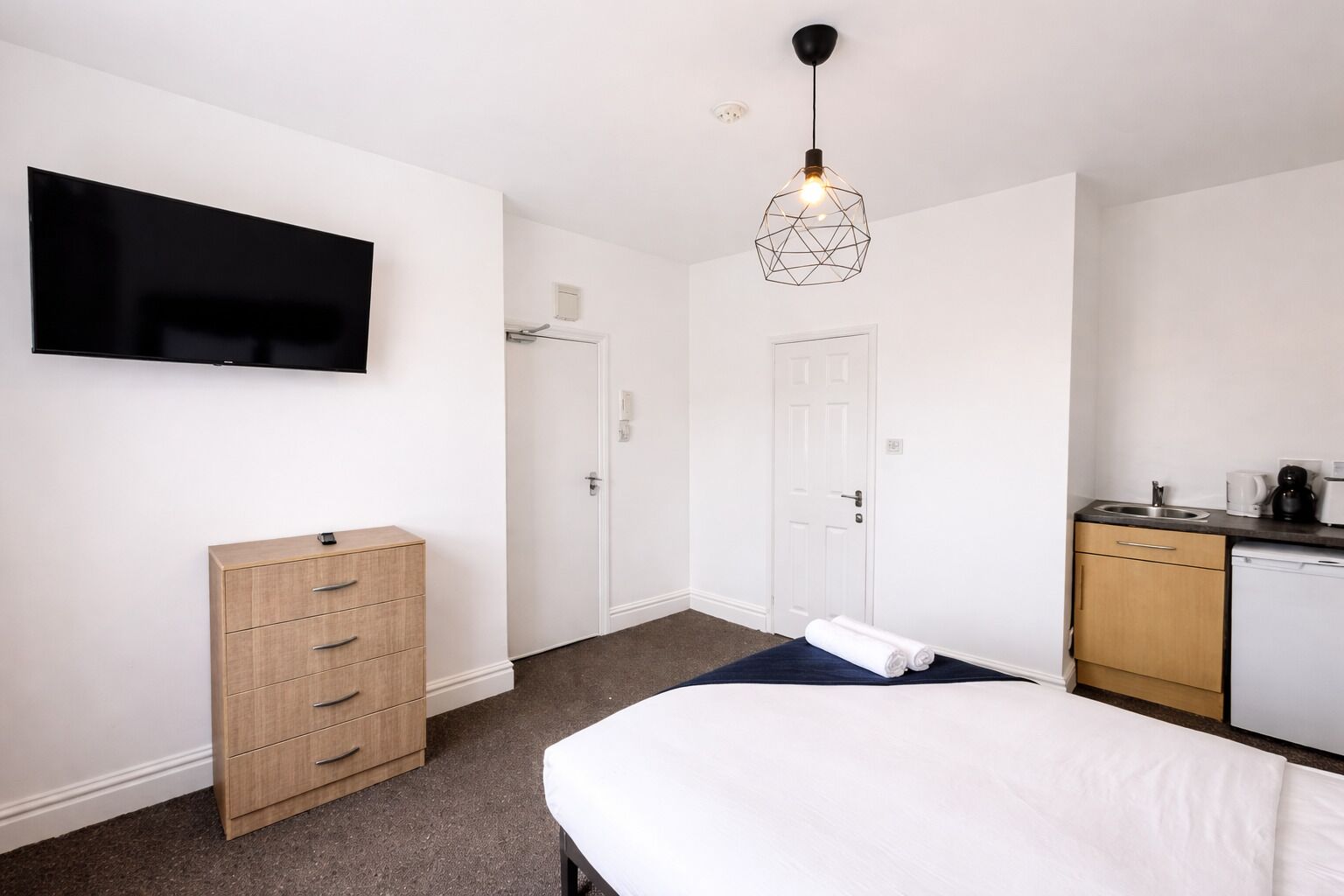 A hotel room with a bed, a TV mounted on the wall, a wooden dresser, a kitchen area with a sink and dishwasher, and a geometric pendant light.