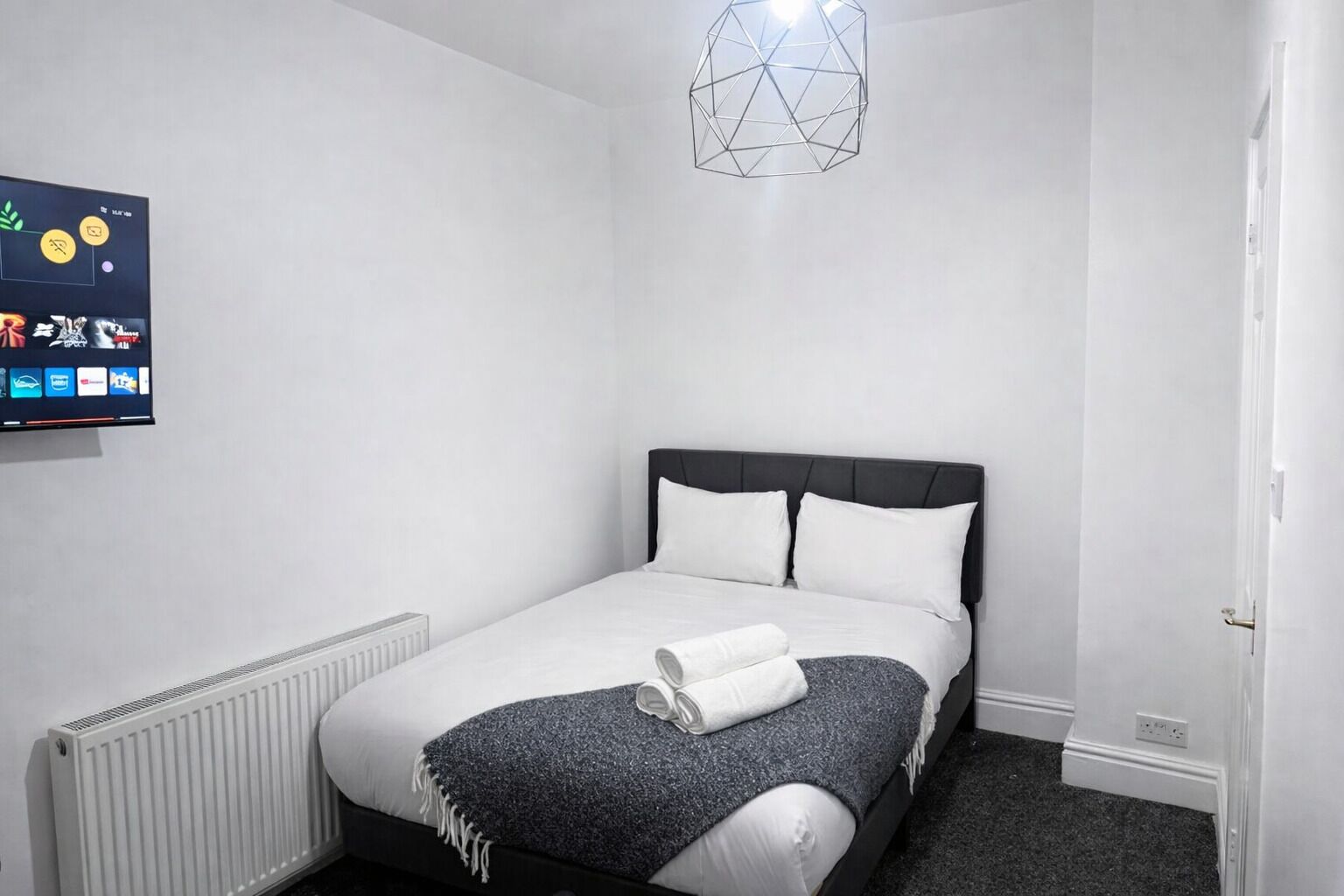 A modern bedroom with a bed, a radiator, a wall-mounted TV, and a geometric light fixture.