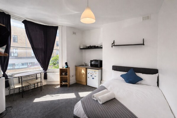 Une chambre d’hôtel de taille réduite, comprenant un lit, un bureau, une kitchenette et une fenêtre avec des rideaux.