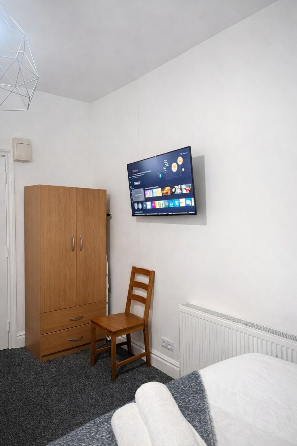 Ein Schlafzimmer mit einem Bett, einem Holzschrank, einem Sessel, einem Heizkörper und einem an der Wand befestigten Fernseher.