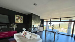 A modern living room with a white sofa, a dining area with a table and chairs, a fireplace, and large windows offering a view of the outdoors. - Guest Room: “Paris Style” with Shared Pool, Wi-Fi, and Air Conditioning (Lumio)