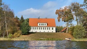 A large white building with a red roof, situated by a calm lake, surrounded by trees and vegetation.
