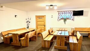 A room with wooden benches and tables, a door labeled 'TOILETA', a radiator, and a TV mounted on the wall.