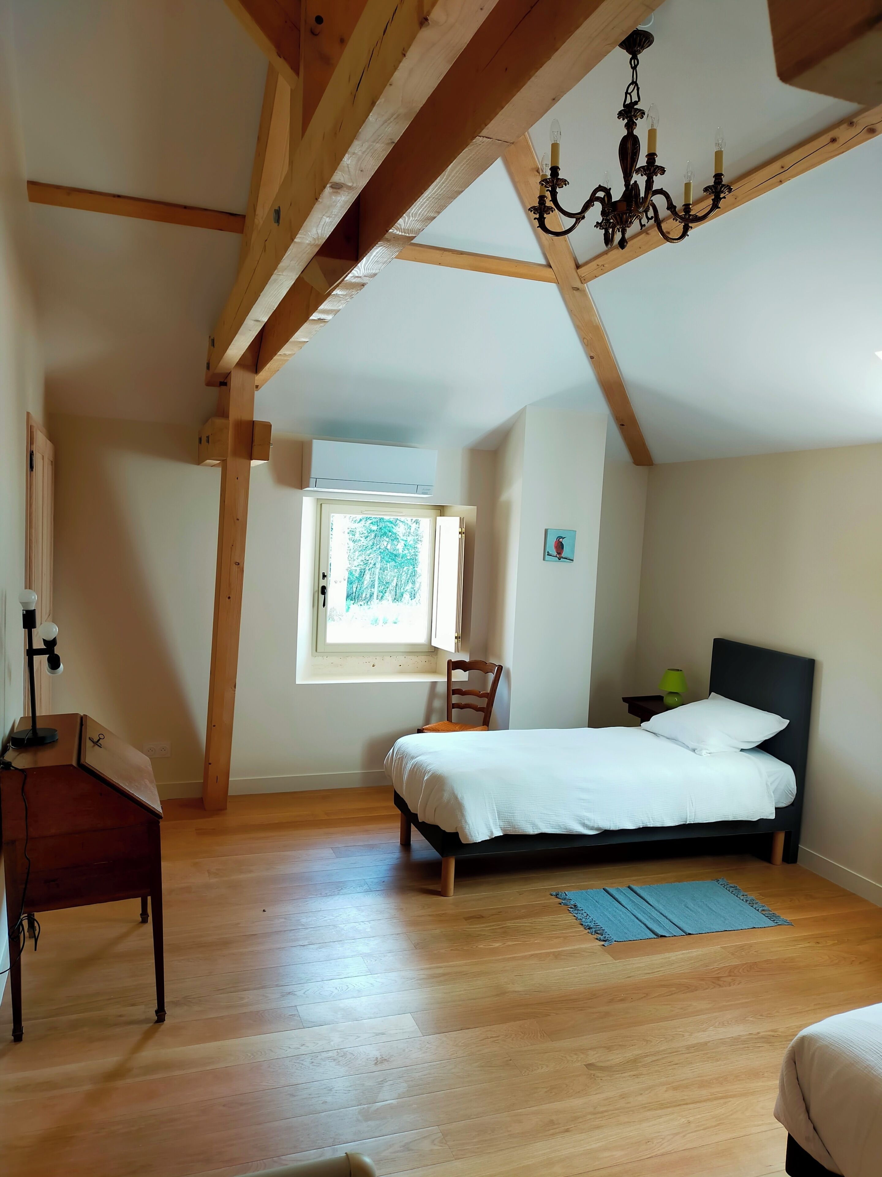 A bedroom with a bed, a chair, a desk, a window, and a chandelier.