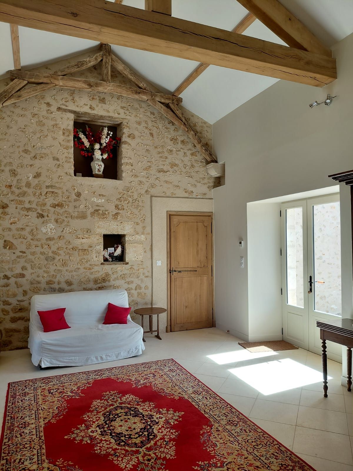 Ein Zimmer mit einer Steinsandsteinwand, Holzbalken, einem weißen Sofa mit roten Kissen, einem gemusterten Teppich und einer Holztür.