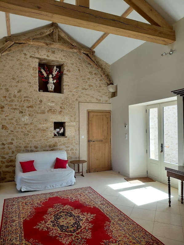 A room with a stone wall, wooden beams, a white sofa with red cushions, a patterned rug, and a wooden door.