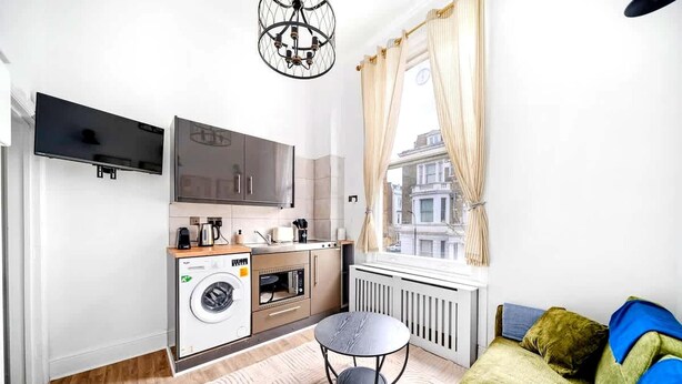 Ein kompakter Wohnbereich mit Küchenzeile, Waschmaschine, Esstisch, Sofa und einem großen Fenster mit Blick auf Gebäude.