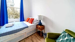 Ein Schlafzimmer mit einem Bett, einem Nachttisch, einer Lampe, einem Fenster mit Vorhängen und Blick auf Gebäude sowie einem grünen Sessel.