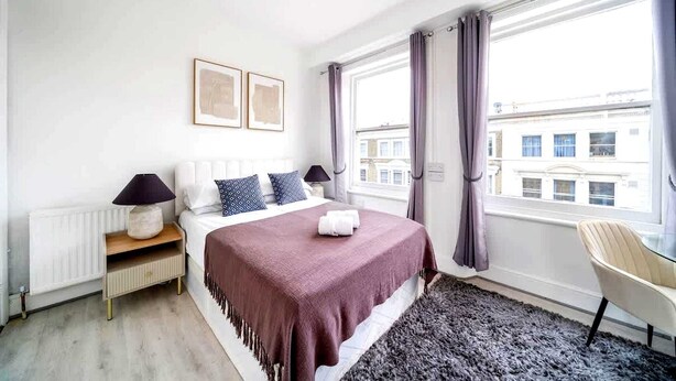 A bedroom with a bed, two bedside lamps, a window with curtains, a chair, and a radiator. - West Kensington Modern Couple Retreat (Greater London)