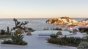 A coastal hotel with a pool, surrounded by cacti and flowers, overlooking the sea and a rocky island.