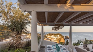 An outdoor dining area with a view of the sea, white pillars, and wicker furniture.